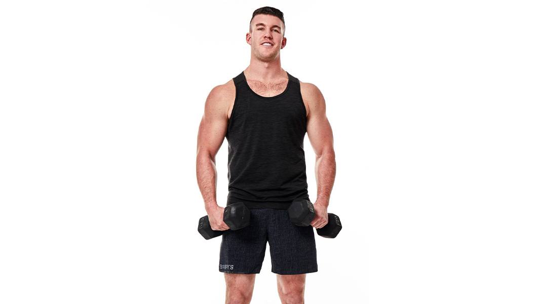 Barry's Bootcamp instructor Connor Miller in black tank top smiling while holding dumbbells down by sides on white background