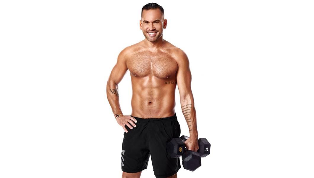Shirtless Barry's Bootcamp instructor Keoni Hudoba with one hand on hip and one hand holding dumbbell on white background