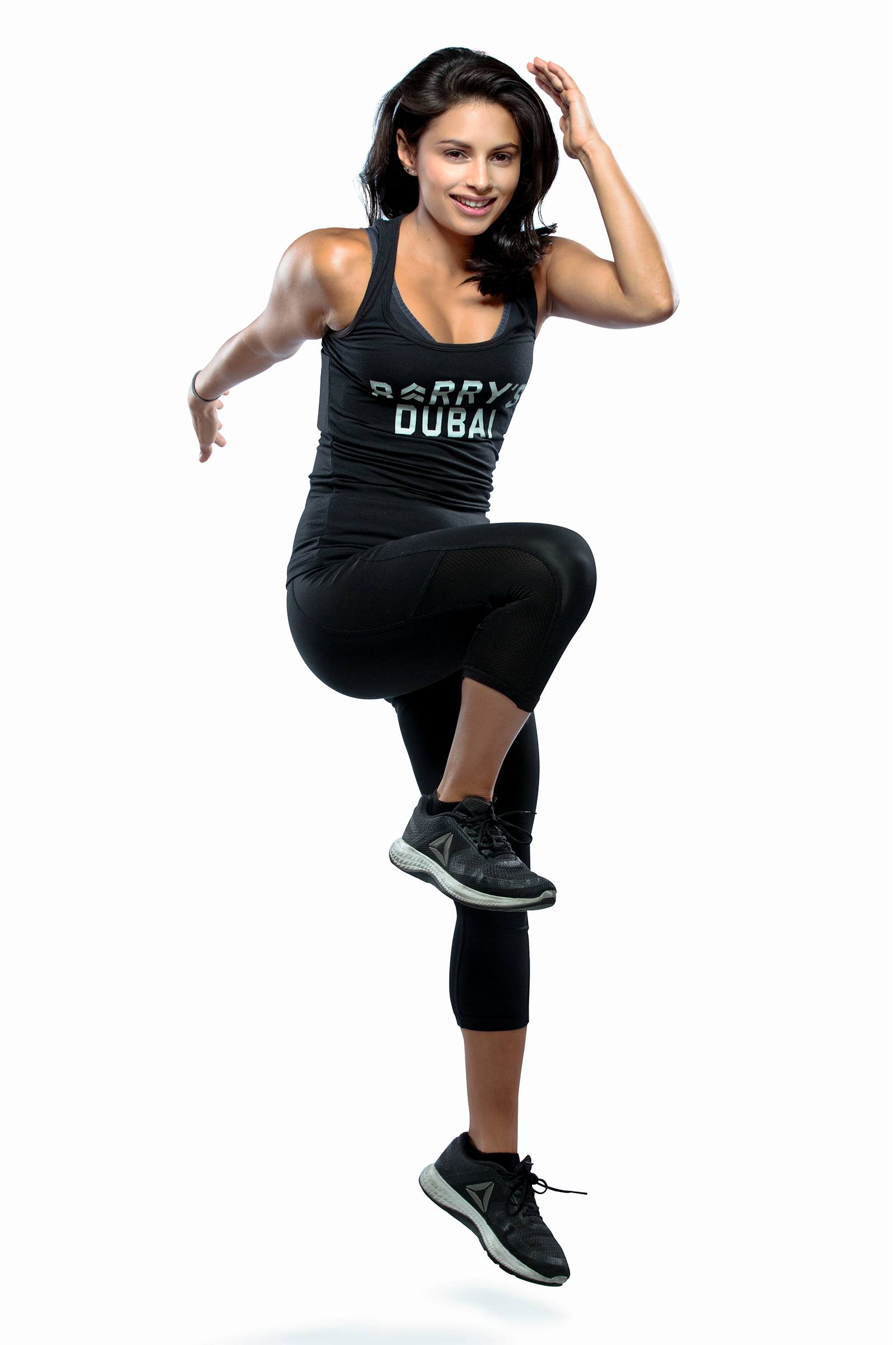 Instructor Nasha Sarkari in black Barry's Bootcamp tank top doing skip exercise move on white background