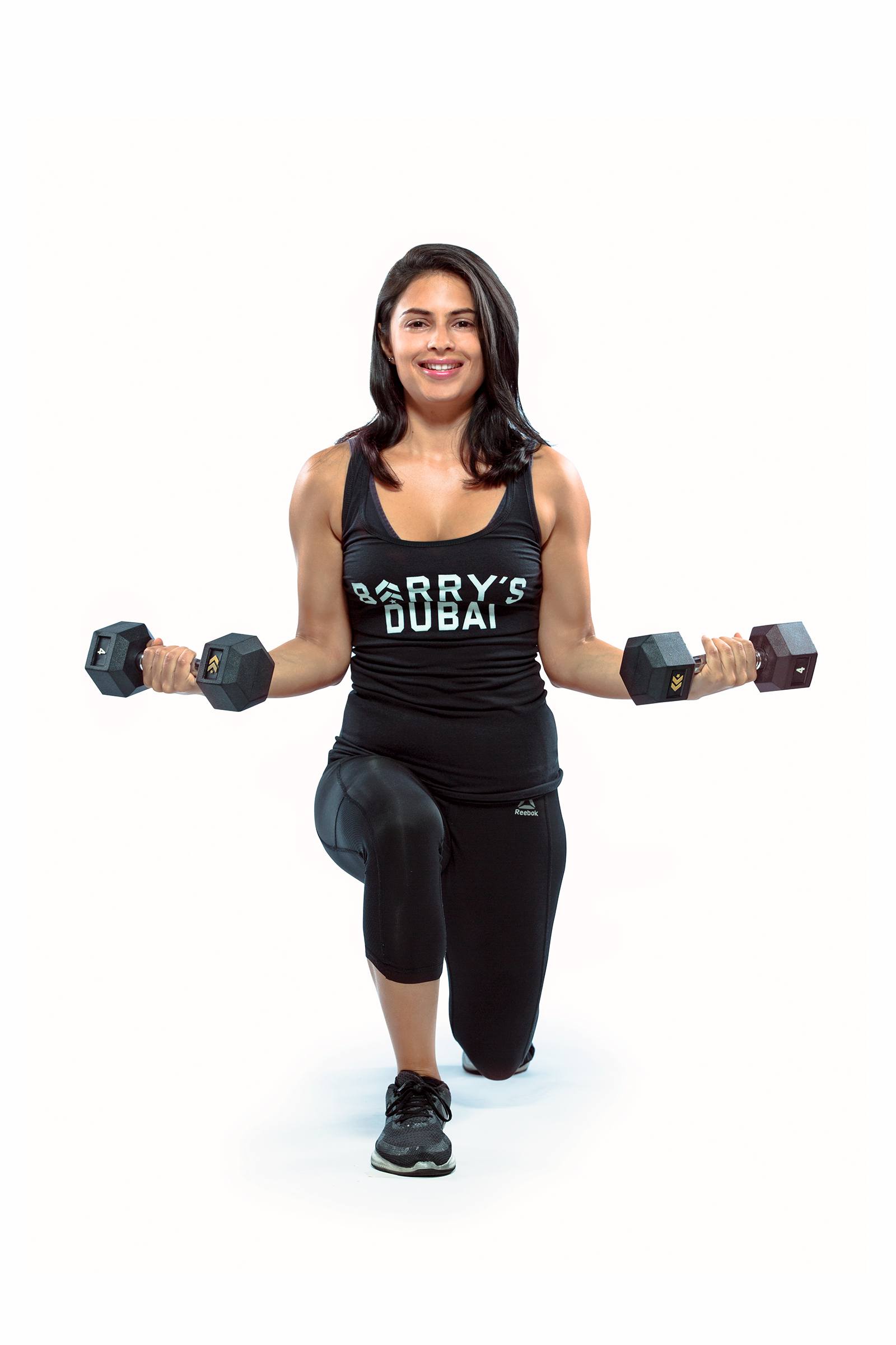 Instructor Nasha Sarkari in black Barry's Bootcamp tank top smiling and holding dumbbells while doing lunge on white background