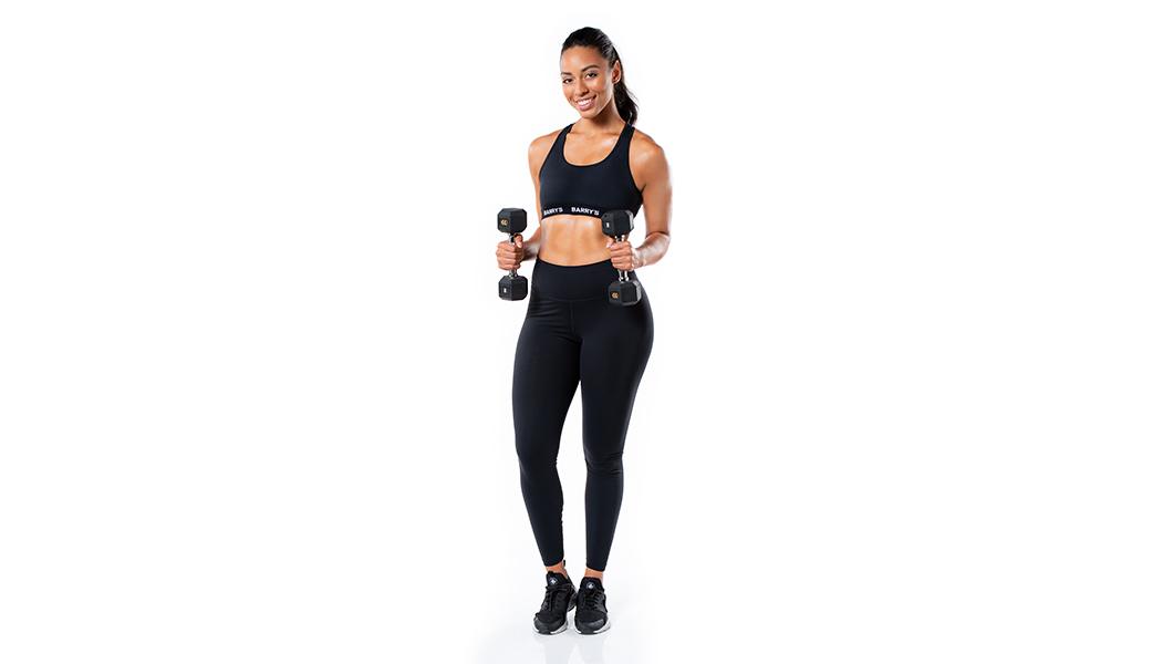 Barry's Bootcamp instructor Mercedes Owens wearing a black sports bra against a white background holding dumbbells vertically in each hand.