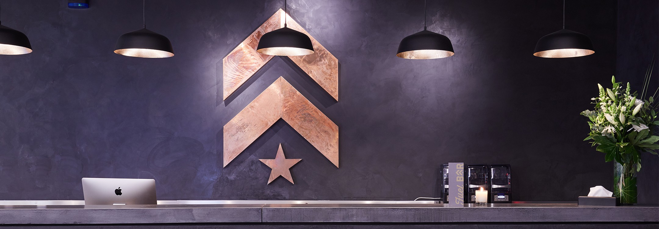 Gold Barry's Bootcamp upward arrows and start logo on dark blue wall behind front desk at Canary Wharf location