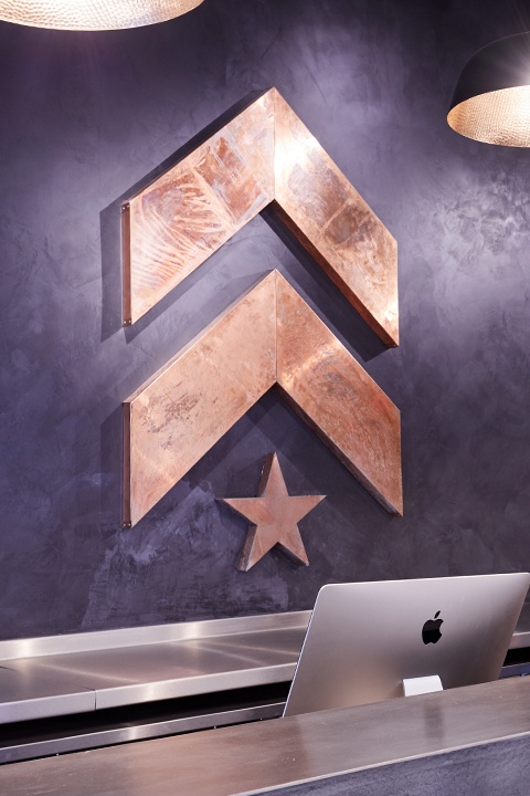 Gold Barry's Bootcamp upward arrows and start logo on dark blue wall behind front desk at Canary Wharf location