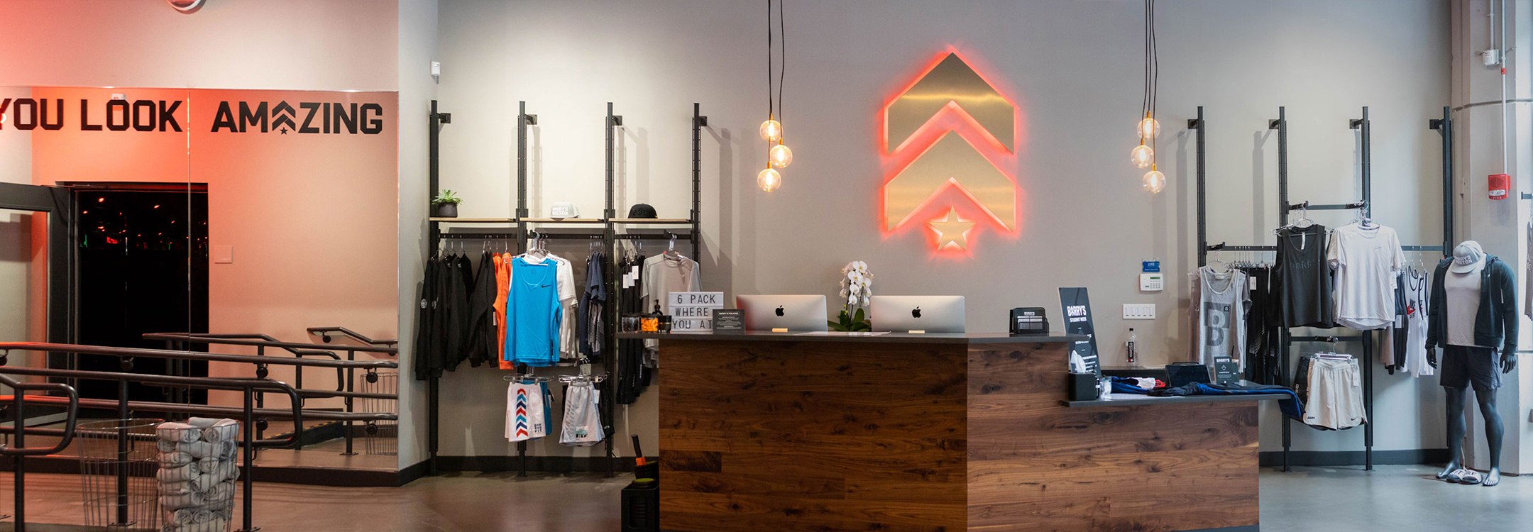 Clothing racks and gold Barry's Bootcamp upward arrows and star logo with red backlighting on wall behind reception desk at Back Bay studio location