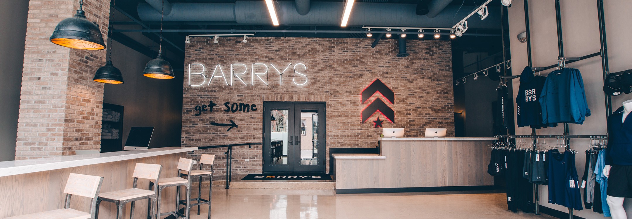 Grey Barry's Bootcamp upward arrows and star logo with red backlighting and White Barry's sign on wall behind reception desk at Lincoln Park studio location