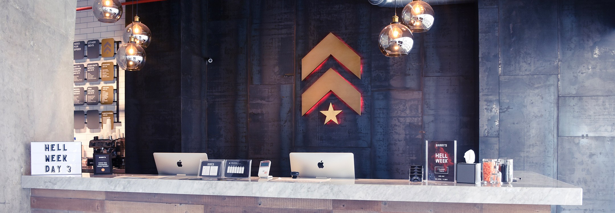 Gold Barry's Bootcamp upward arrows and star logo with red backlighting on grey wall behind reception desk at Dubai Marina studio location