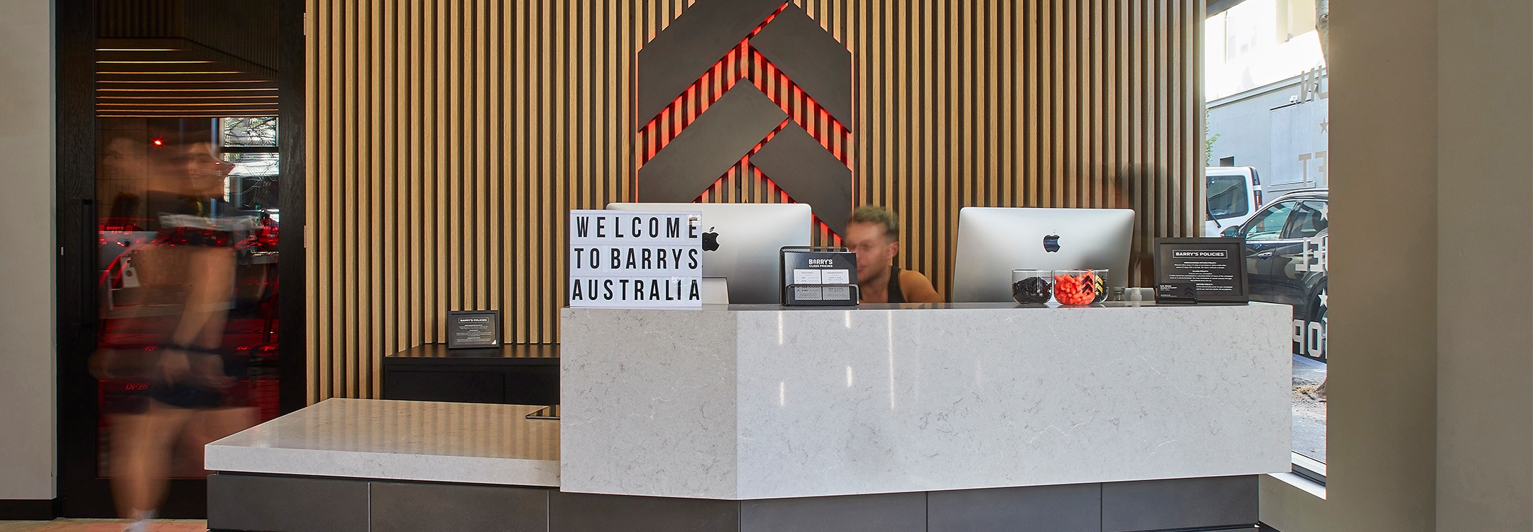 Slate Barry's Bootcamp upward arrows and star logo with red backlighting on wood paneled wall behind desk and sign in Australia studio reception area