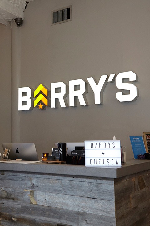 Barry's Bootcamp white sign and yellow and orange logo on wall behind reception desk at Chelsea studio location