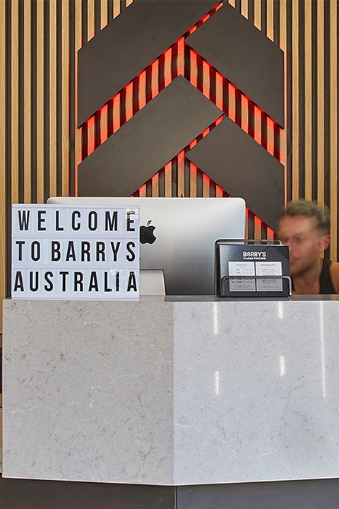 Slate Barry's Bootcamp upward arrows and star logo with red backlighting on wood paneled wall behind desk and sign in Australia studio reception area