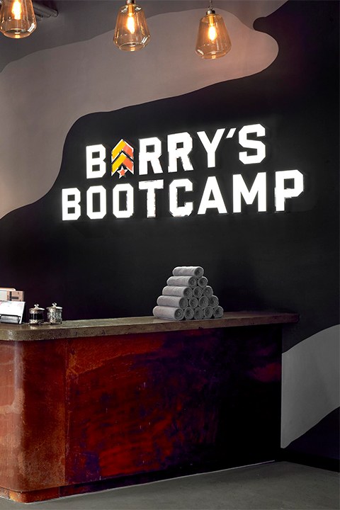 Barry's Bootcamp white sign and yellow and orange logo on wall behind reception desk at London East studio location