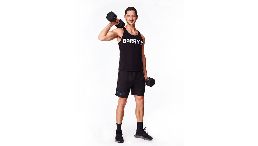 Barry’s instructor wearing a black top standing holding weights confidently smiling against a white background