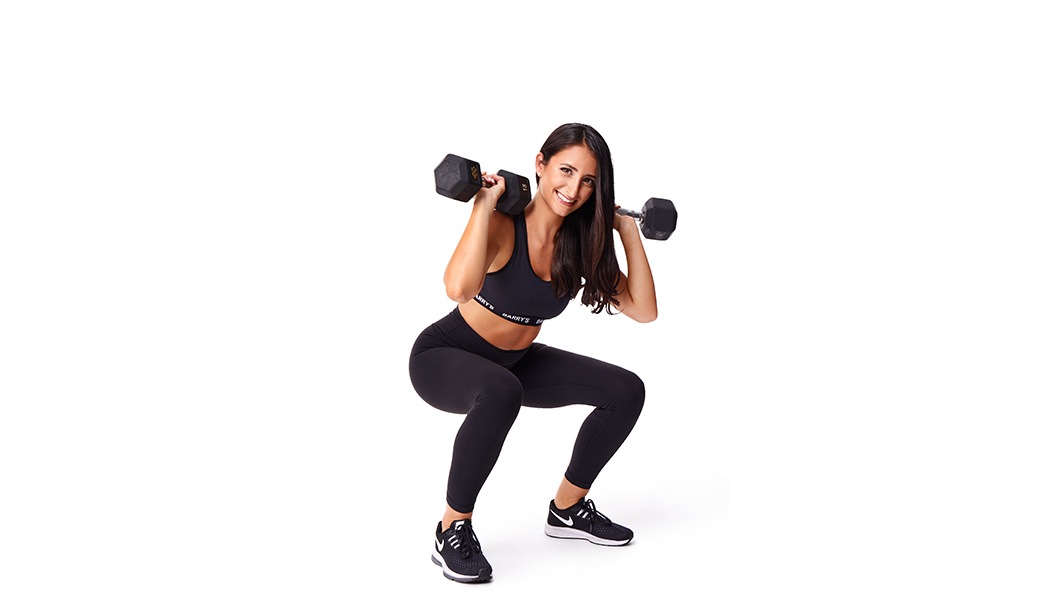 Barry’s instructor wearing a black top standing confidently smiling against a white background squatting with weights on shoulder