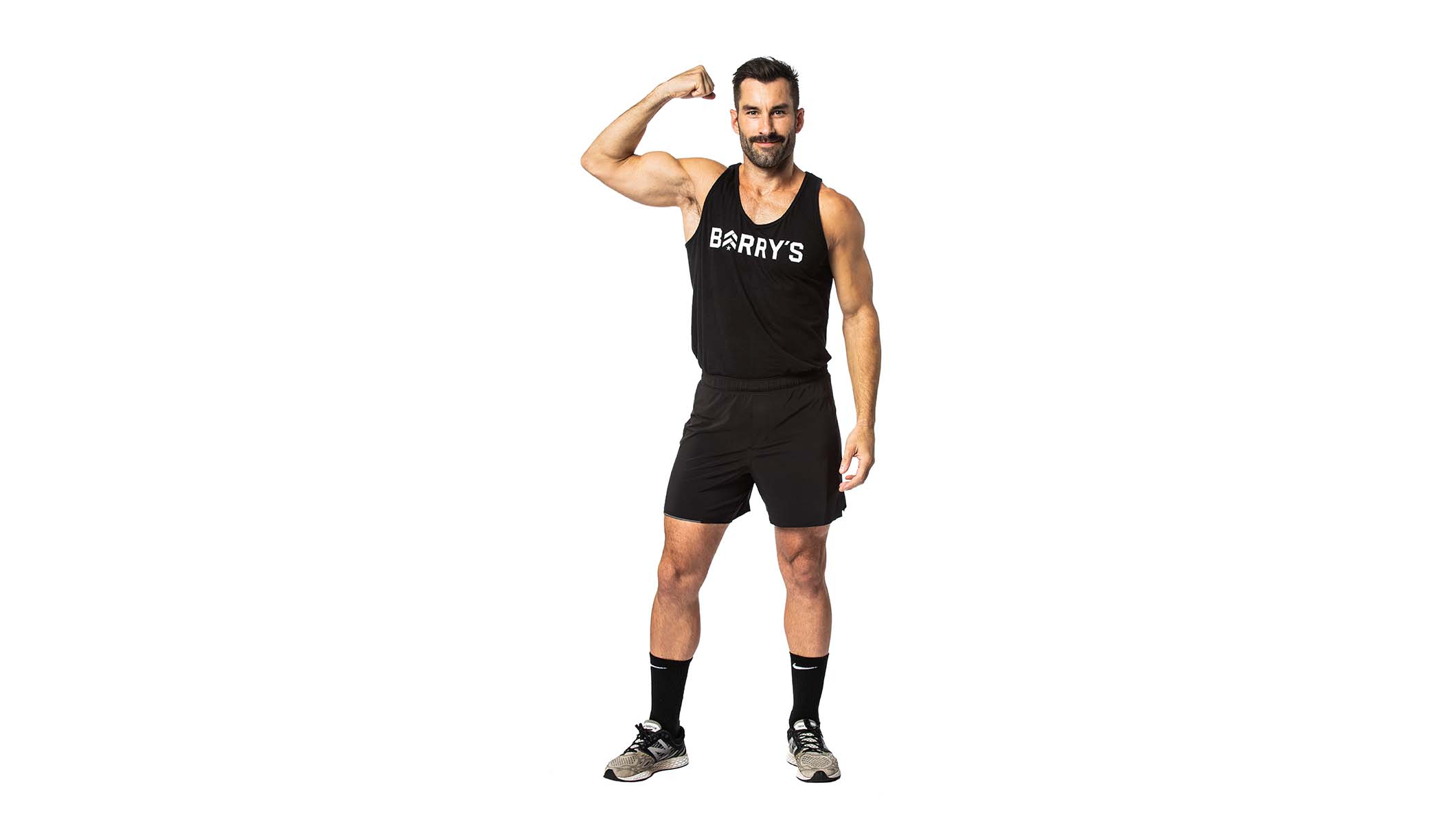Barry’s instructor wearing a black top flexing his bicep standing confidently smiling against a white background