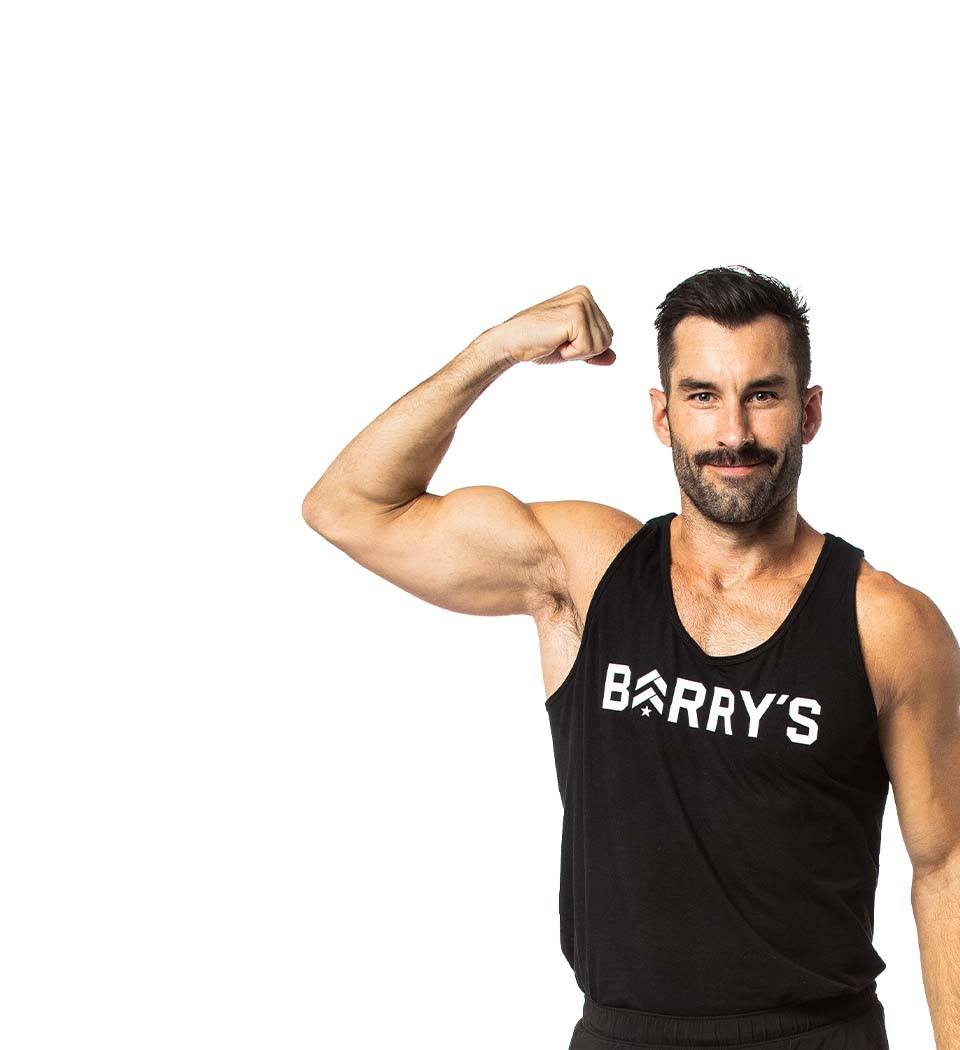 Barry’s instructor wearing a black top flexing his bicep standing confidently smiling against a white background