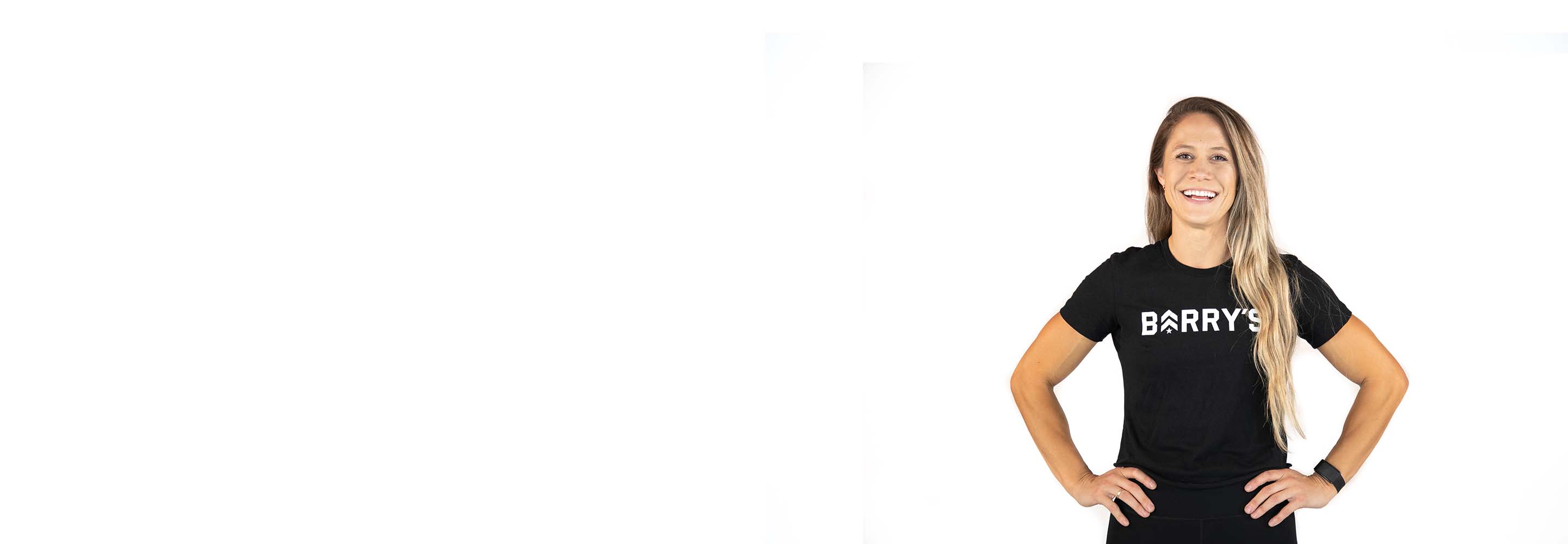 Barry's instructor standing confidently with hands on hips, wearing a black top against a white background, ready for the next workout challenge.