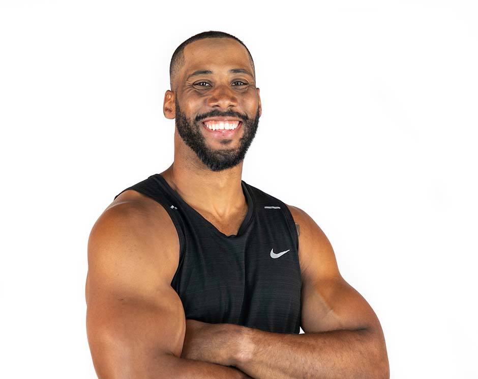 Corey Lemonier instructor standing confidently with arms crossed in front, wearing a black tank top against a white background, ready for the next workout challenge.