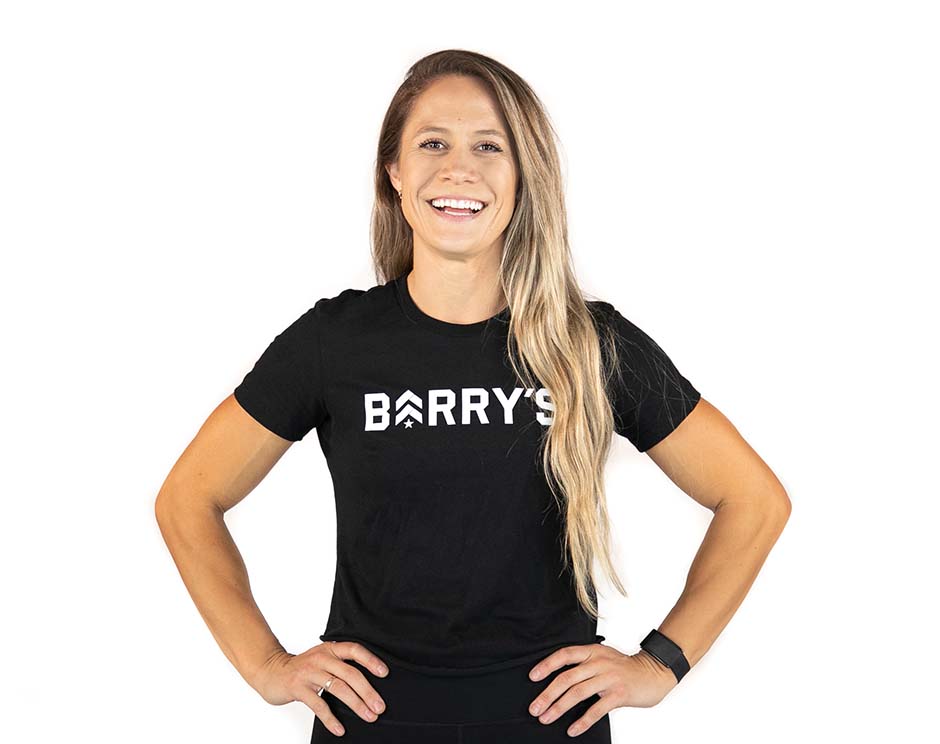 Barry's instructor standing confidently with hands on hips, wearing a black tank top against a white background, ready for the next workout challenge.